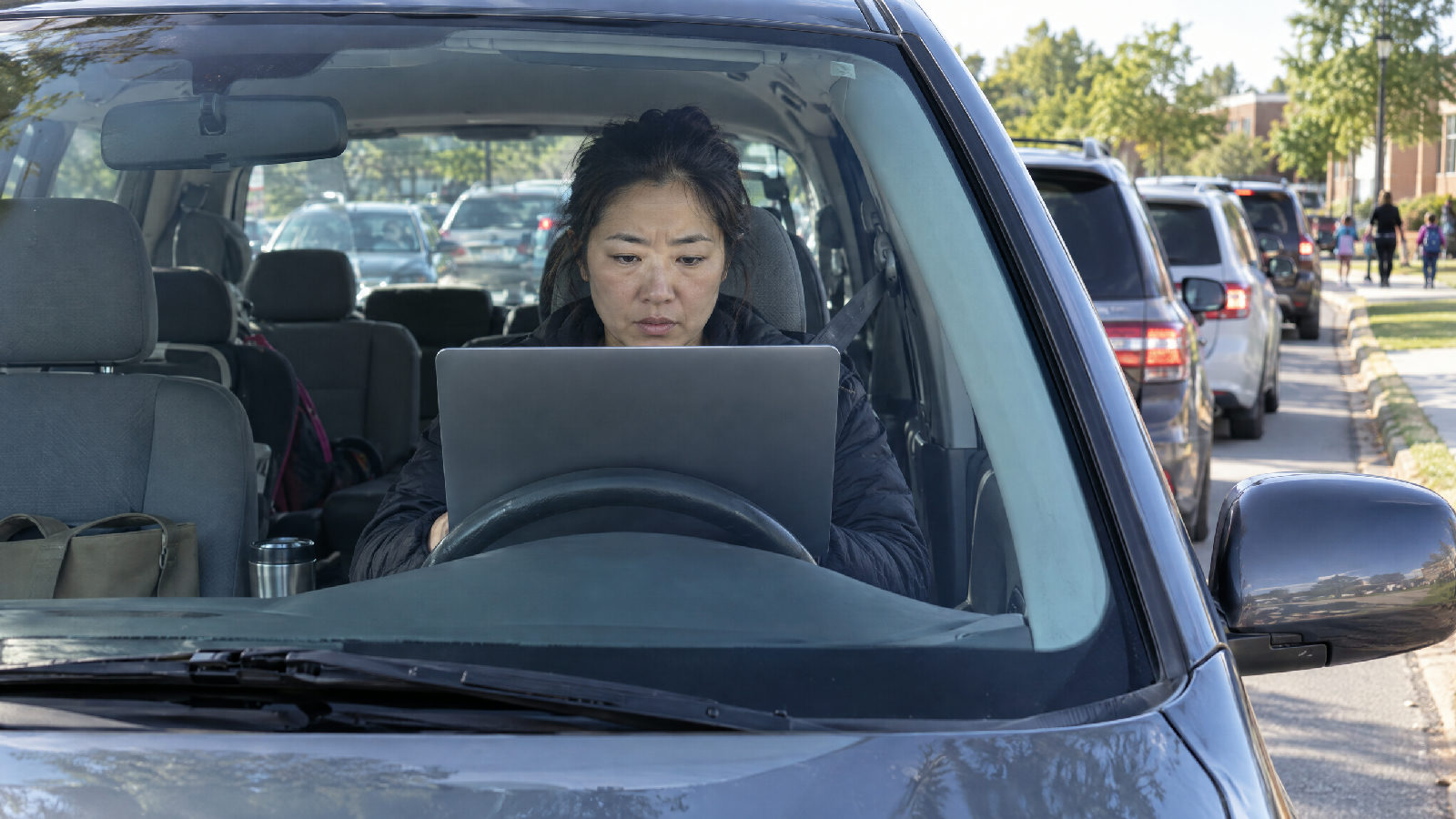 A treasurer working on a laptop propped on her steering wheel while she waits in a school pickup line