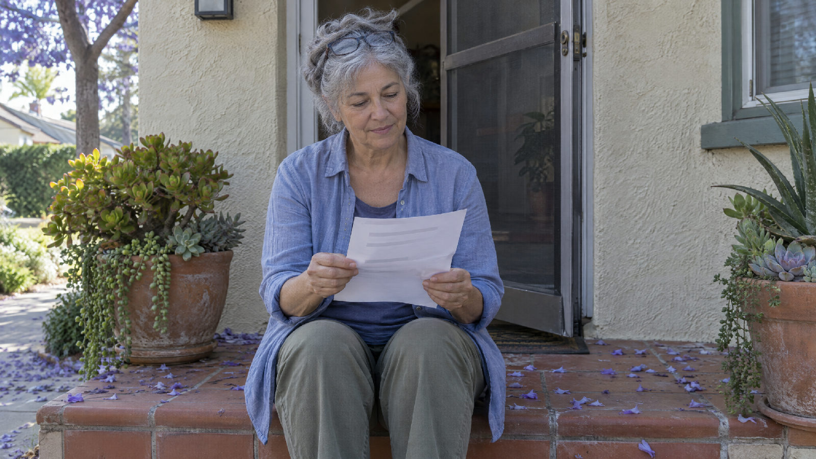 A homeowner on her front step in morning light unfolding a single sheet she found tucked in her screen door, jacaranda petals scattered on the step