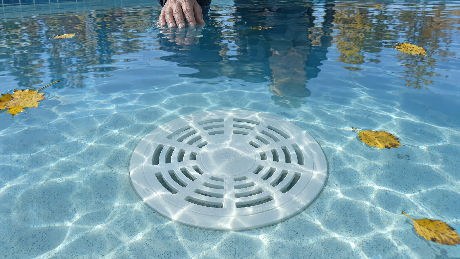 Seen from just above the water of a closed community pool on a clear autumn morning, the round drain cover large and centred on the pool floor, a board member's hand at the top edge, leaves floating