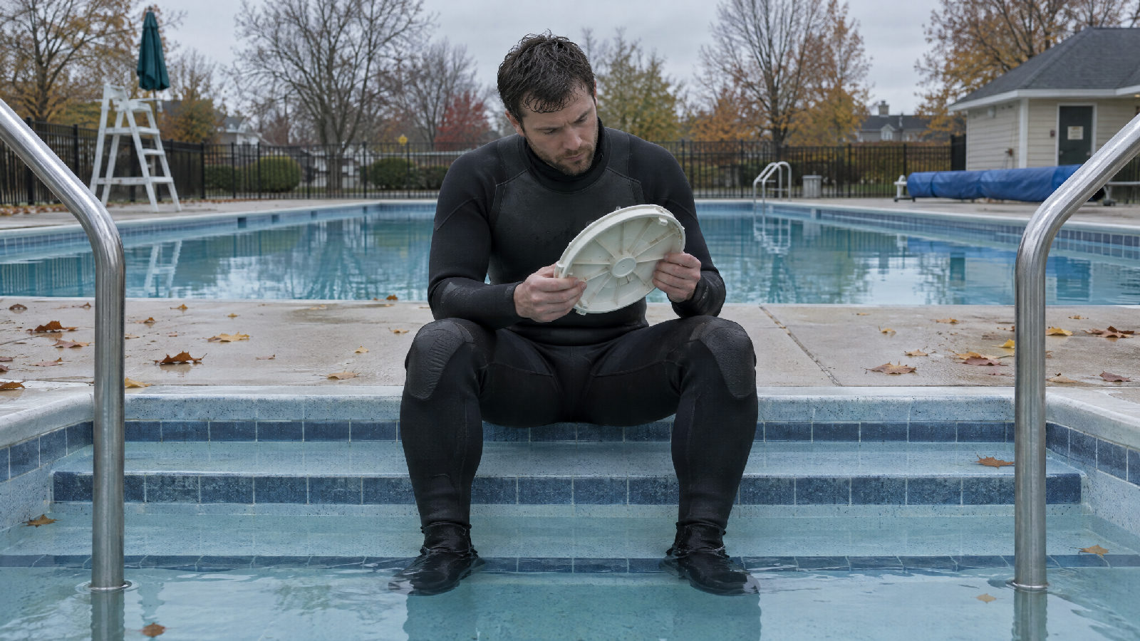A pool technician in a wetsuit sitting on the steps of a closed community pool, holding a round white drain cover in both hands and turning it over to inspect its underside