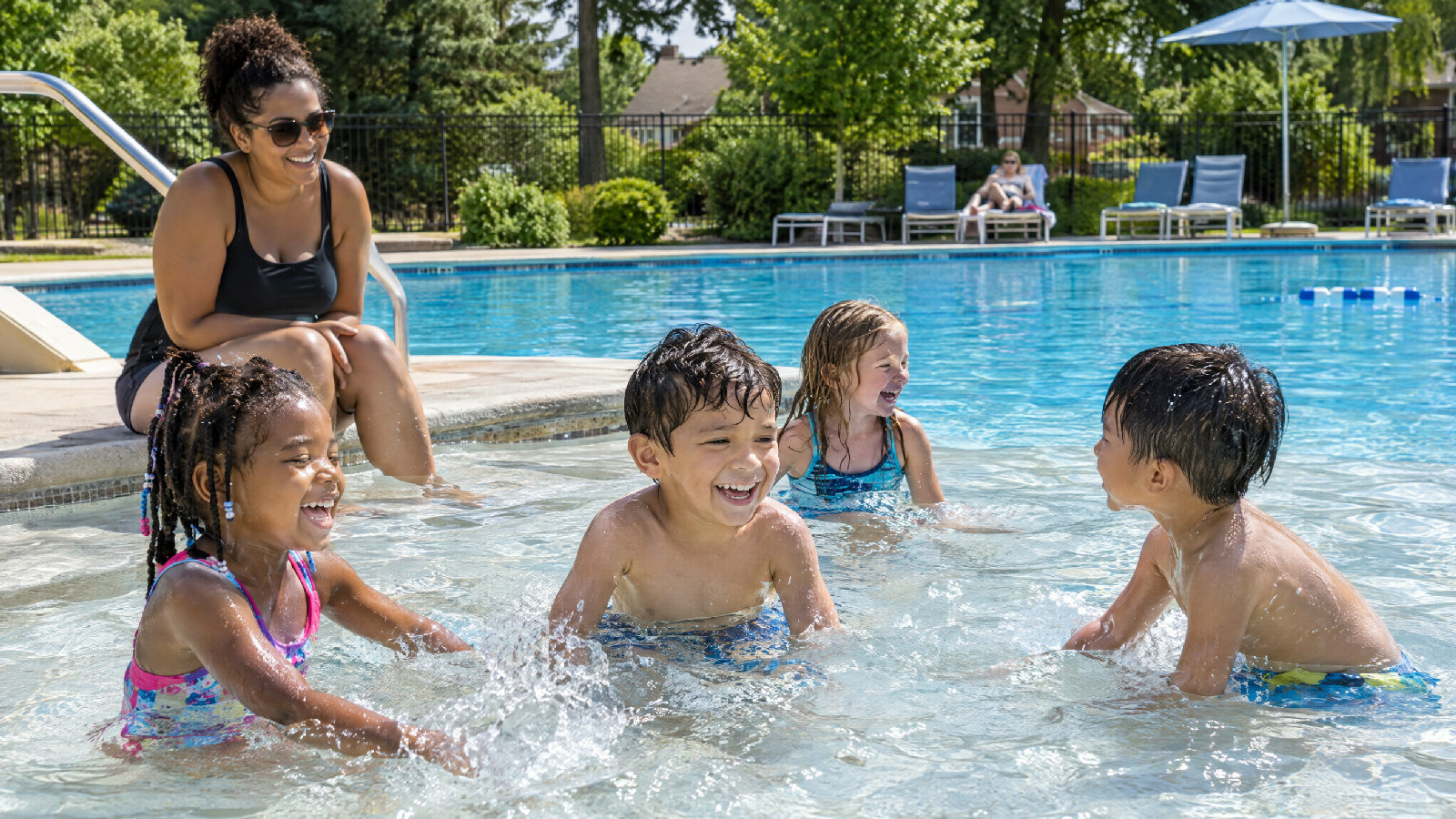 Children splashing in the shallow end of a busy neighborhood pool on a bright summer afternoon while a parent watches from the pool steps, the deep end calm behind them