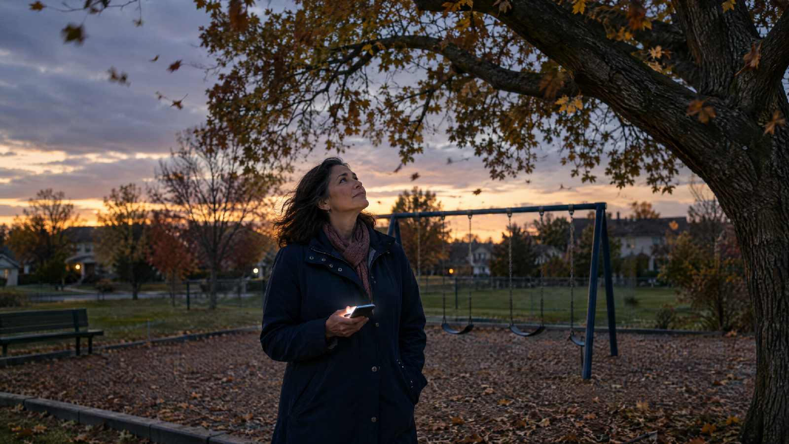 A board member at the edge of an empty playground on a windy autumn evening, phone glowing in her hand, looking up at a large oak leaning over the swings
