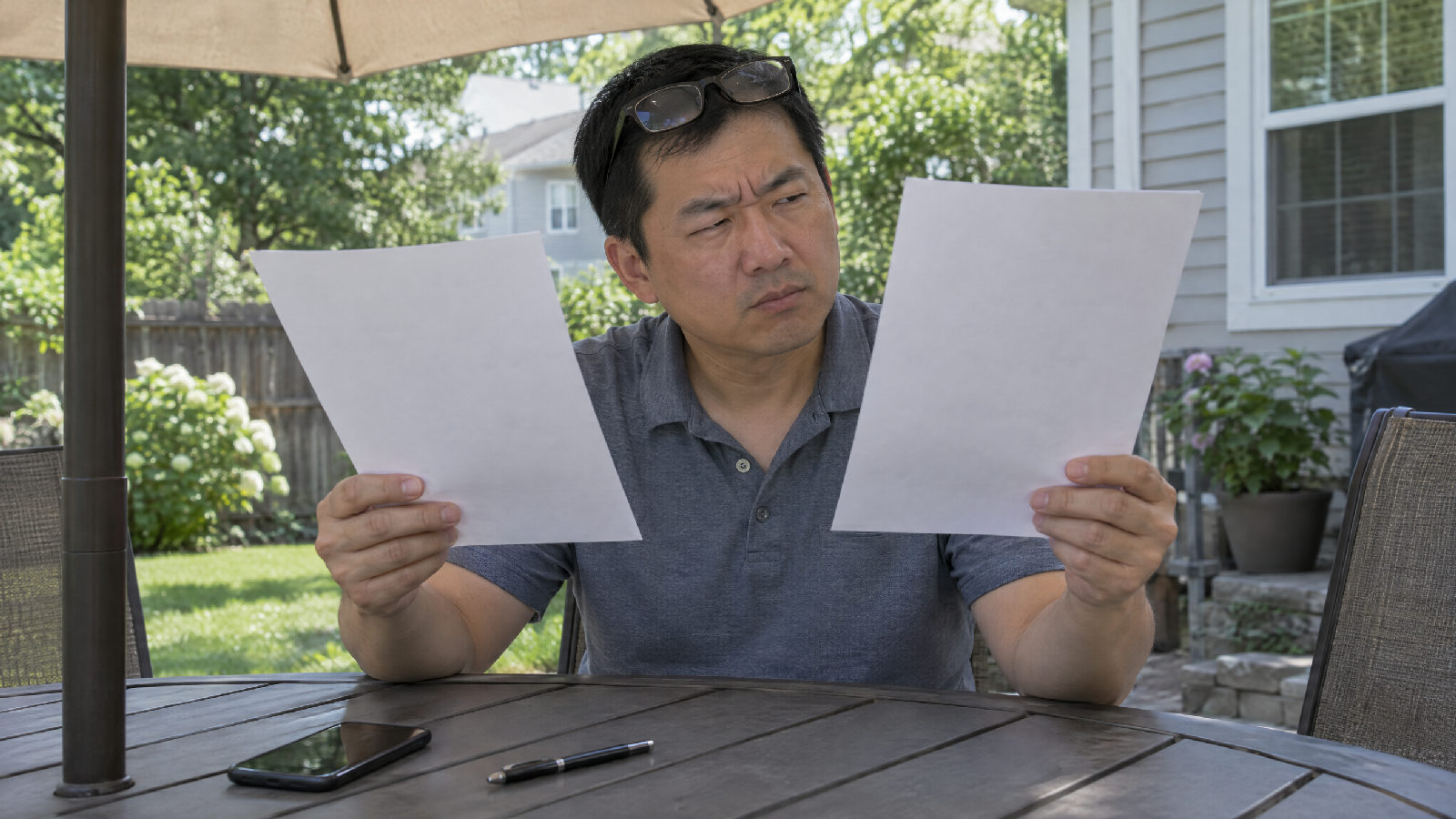 A treasurer at a backyard patio table holding two sheets of paper side by side, comparing them with a skeptical squint