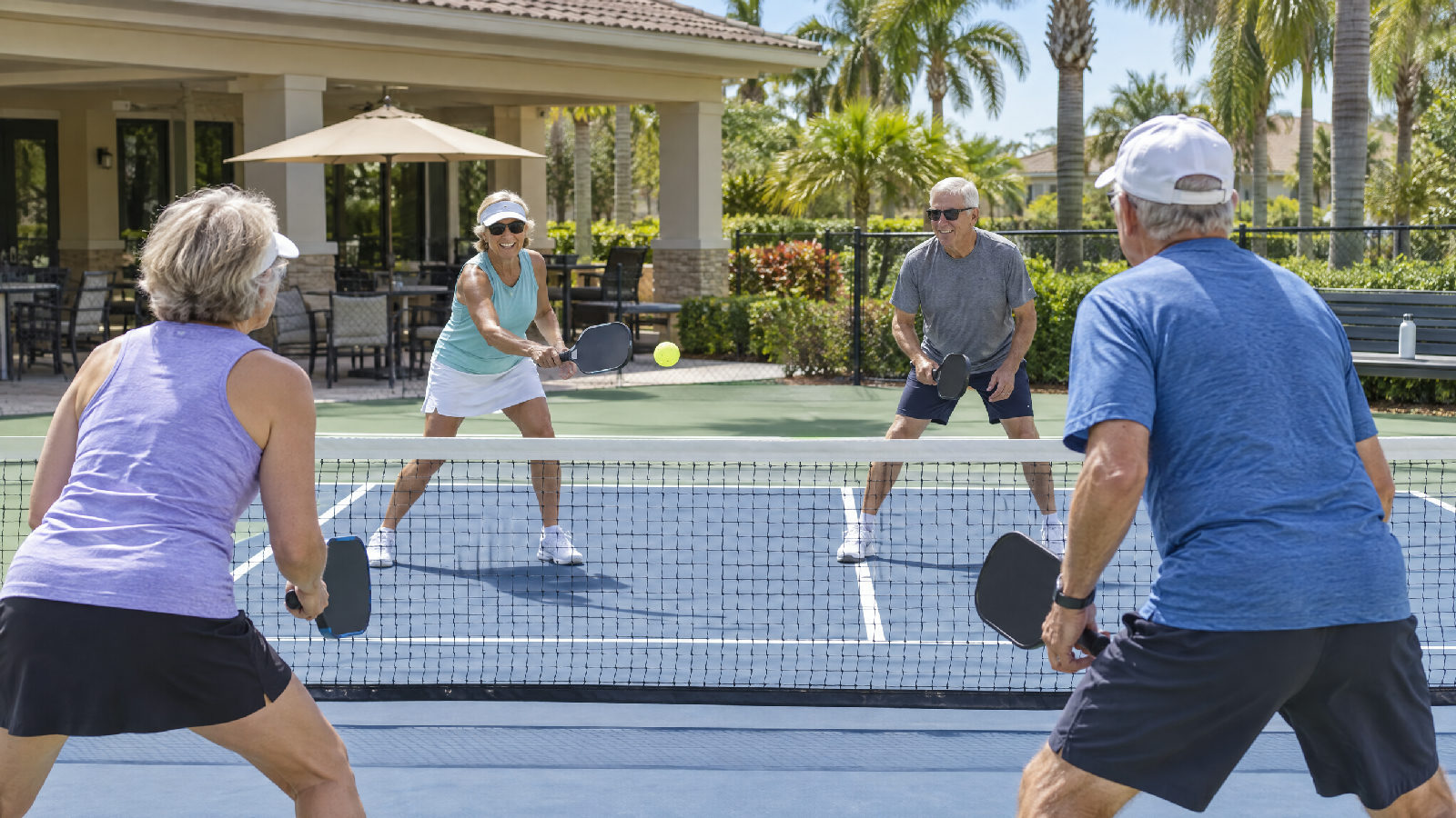 **Paid-off homes, bigger amenities.** The Census Bureau's own guess at why owners without a mortgage pay more. Retirees playing doubles pickleball on a community clubhouse court in late morning, a shaded patio and palm trees behind them