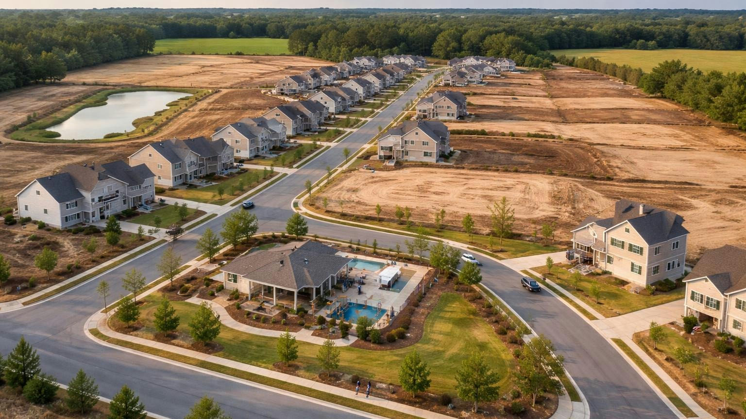 **Built in phases.** The builder runs the association until enough homes are sold. A wide aerial view of a new subdivision at the edge of farmland, half the lots still bare dirt and half finished houses, a retention pond and a small amenity center near the entrance; no readable text